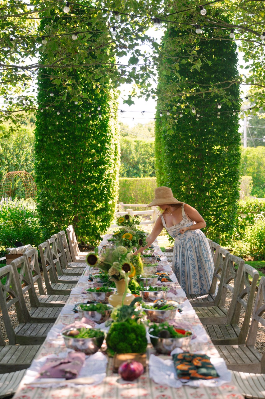 Event planner styling a long outdoor communal dining table with fresh produce, floral centerpieces, and place settings for the Agua by Agua Bendita x CAKE brand activation in the Hamptons.